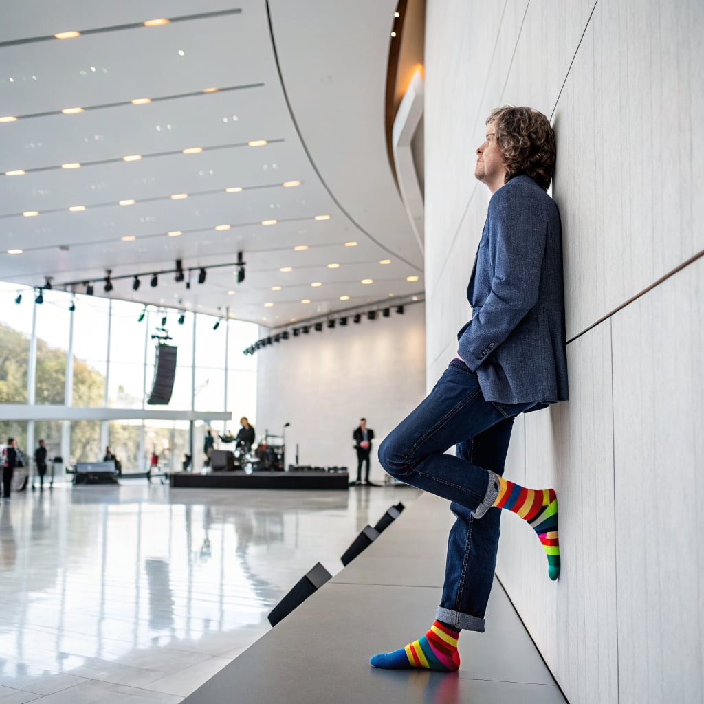 A man in a blazer and colorful socks leans against a wall in a modern event space.
