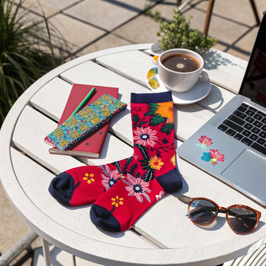 A pair of colorful floral socks is displayed on a table alongside a notebook, a cup of coffee, a laptop, and sunglasses.