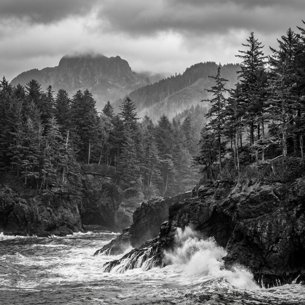 A black and white image of rocky coastline with waves crashing against the shore and mountains in the background.